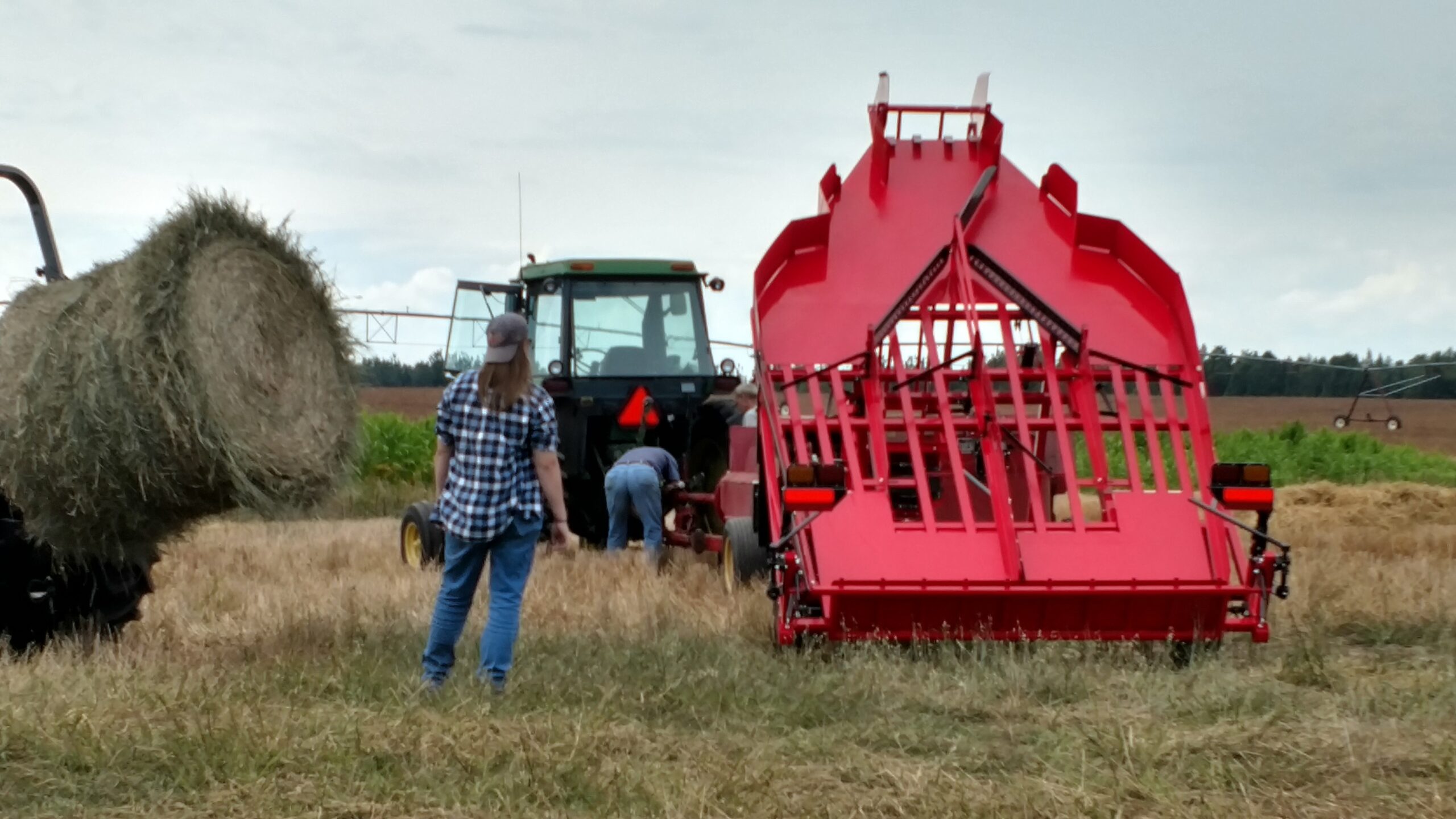 Farm workers operating machinery in a field, featuring a red hay baler and a green tractor, with hay bales in the background.