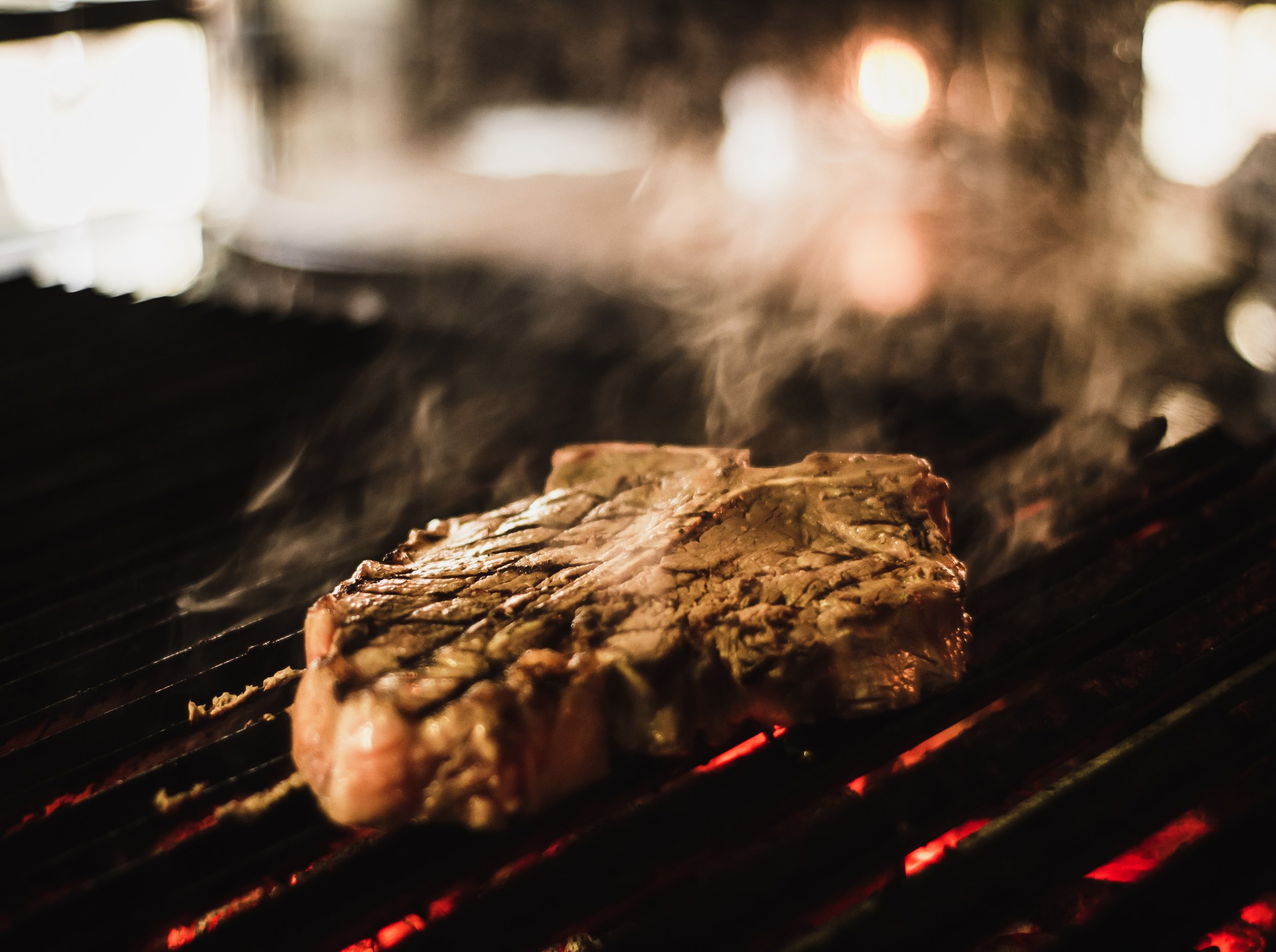 Grilled steak sizzling on a barbecue with smoke rising, showcasing the charred grill marks and juicy texture against a backdrop of glowing embers. Perfect for food enthusiasts and grilling recipes.